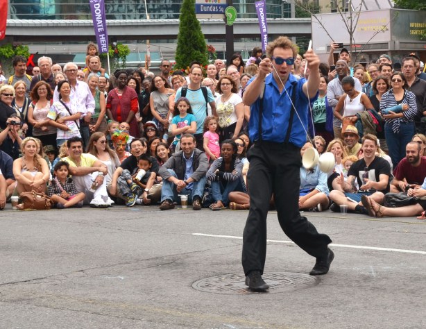 A street performer is using a diabolo to entertain a crowd of people who are sitting and standing on the sidewalk around him.