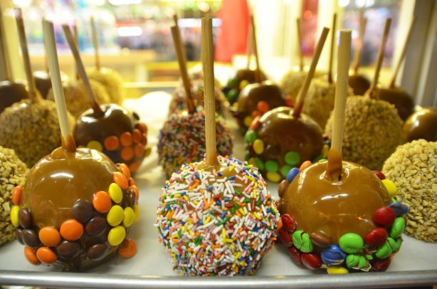 A display of candy apples for sale.  Some are coated with smarties candies, some with multi coloured sprinkles and some with crushed nuts. 