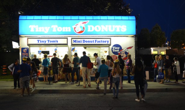 Evening shot as the sky is getting darker.  Tiny Tom donut stand all lit up, with a line up of people waiting to buy donuts. 