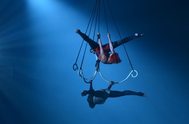 Two acrobatic women hanging from a large triple hook shaped bar suspended from the ceiling.  Bathed in blue light. 