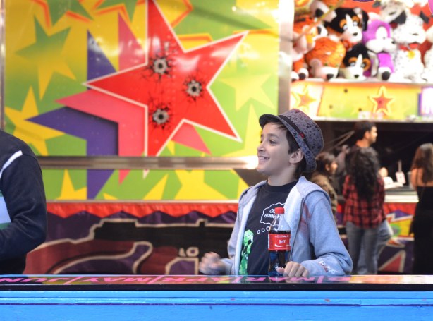 A boy wearing a fedora is standing beside one of the midway games.  He is holding a coke bottle with the name Jeff on it.  Behind him is a large picture of 3 stars. 