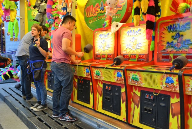 Two young men are hammering away at Whac-a-mole while a woman who is running the game is talking to the people who are watching 