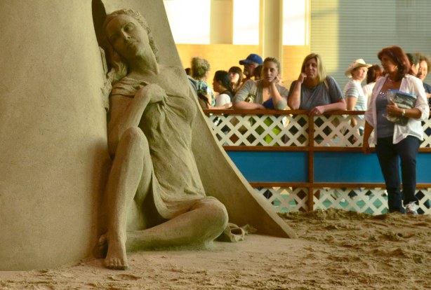 Sand sculpture of a woman sitting on the ground, looking wistfully through a round window.  In the background are some woman standing behind a wood barricade who are looking at the sand sculpture