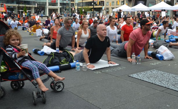 Group of people at Dundas Square doing yoga postures as part of a yogathon.  A young girl sits in a stroller with a Starbucks cup in her hand.  She is watching the yoga (but smiling at the camera)
