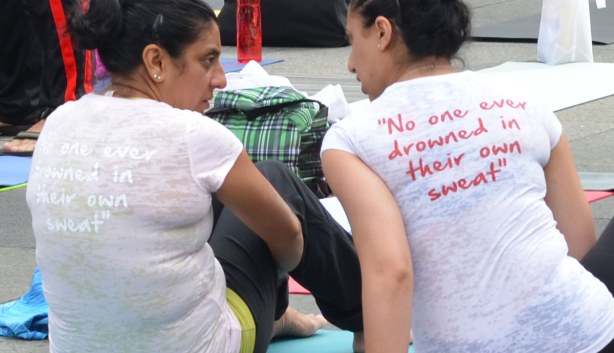 Two women are sitting on the ground and talking to each other.   Their backs are to the camera.  On the back of their Tshirts are the words 'no one ever drowned in their own sweat'.