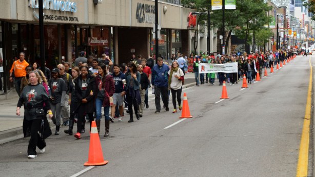 Looking south down Yonge Steet.  A large group of people are walking as part of the AIDS walk.  They are in the far lane of the street.  Orange traffic cones run down the middle of the street to protect the walkers from traffic. 