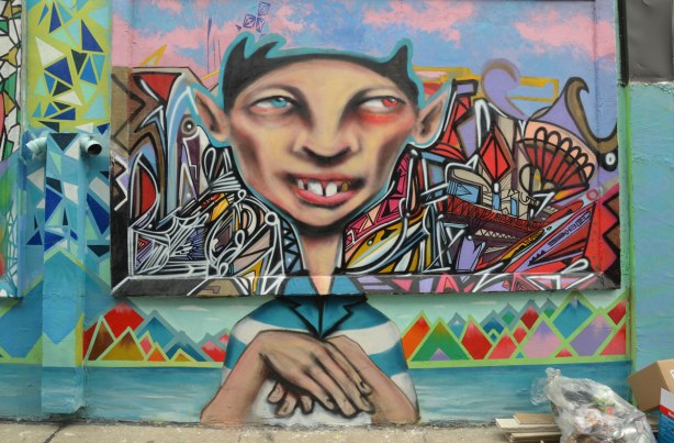 Colourful graffiti in a Kensington lane showing a person with a large head, from the ribs up. Hands are crossed in front of him. Multicoloured geometric shapes are on either side of his head.