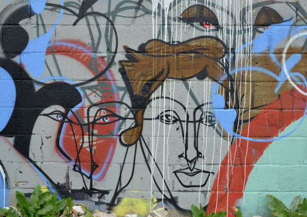Street art painting on the side of a brick wall, black line drawn faces with some reds, whites and blues , and one with brown hair 