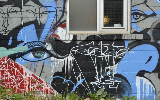 Street art painting on the side of a brick wall, black line drawn faces with some reds, whites and blues , and two realistic looking eyes on either side of a window 
