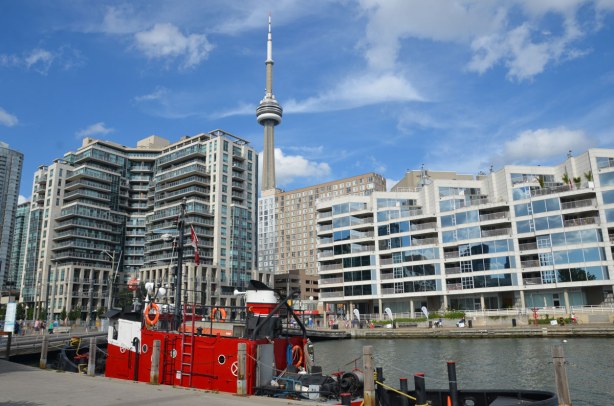 A bright red tugboat is tied up in the foreground. Many glass windows of many condos are in the photo. The top of the CN tower is peaking up over the top of the condos on Toronto's waterfront.