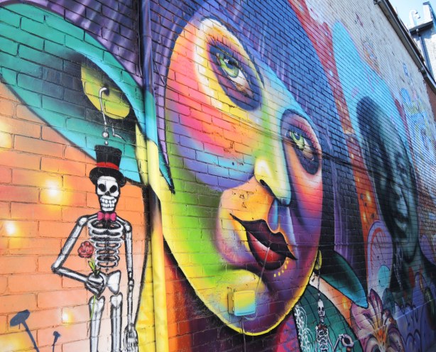 part of a large mural on the side of a 2 storey building, a large multicoloured woman's face. She is wearing earrings that look like skeletons