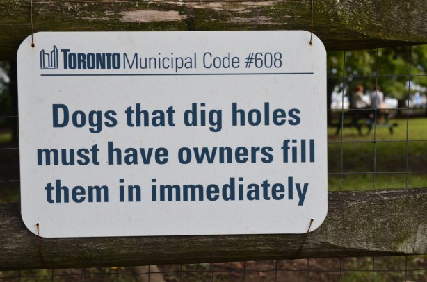 Sign on a wood fence by a park that says, Dogs that dig holes must have owners fill them in immediately.