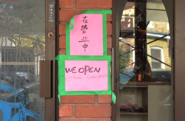 A pink sign that has been taped with green tape beside the entrance of a shop that says We open.  Please come in. 