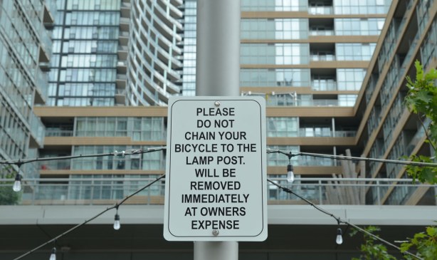 Sign by a sidewalk that says Please do not chain your bicycle to the lamp post.  Will be removed immediately at owner's expense.  Large condo towers are behind the sign. 