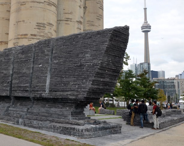 view of Ireland Park from the waterfront, and looking slightly north east. A large shape made of limestone is on the left of the photo and a green space is beside it. The silos of the Canada Malting Company are visible as is part of the Toronto skyline in the distance.