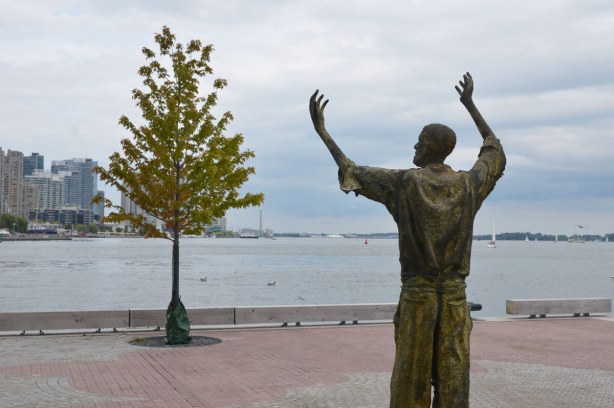 Sculpture of a man with his arms upraised. He is looking over part of the harbour towards downtown Toronto.