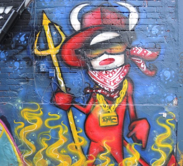 Graffiti painted on a wall that looks like a stylized dog as a red devil with horns, wearing sunglasses and holding a yellow devil's fork
