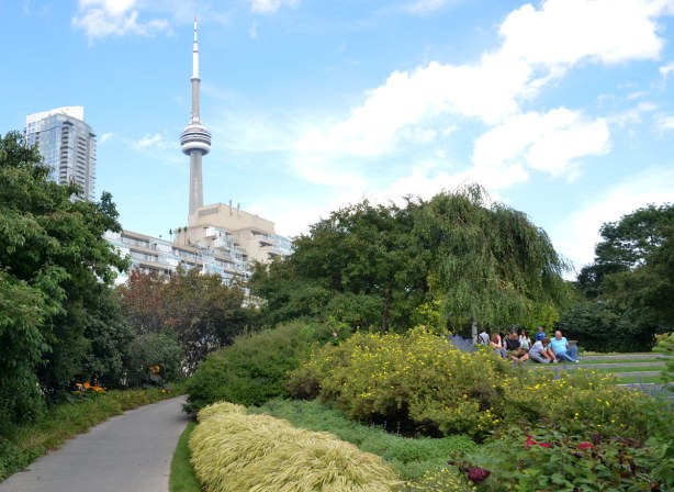 A path leads through a garden, there is a group of people sitting in the park. The CN Tower is in the distance.
