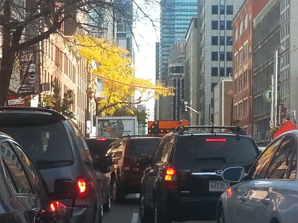 Cars trying to merge from three lanes to one.  Large sign with arrow pointing left.  Tall buildings on either side of the street.  Yellow leaves on the trees because it is October.  