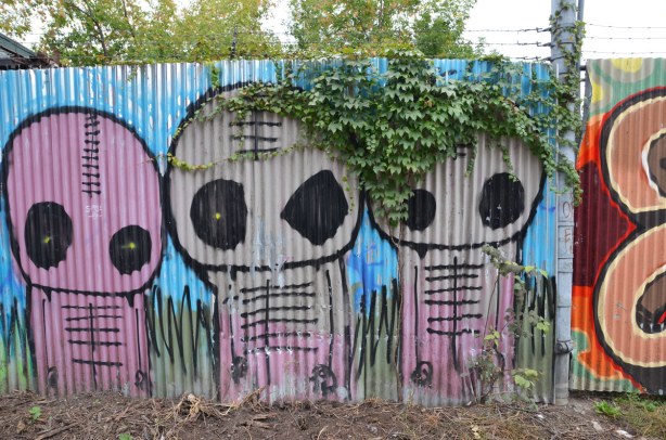 Three large pinkish alien like creatures on a corrugated metal fence.  Some ivy is hanging over the fence