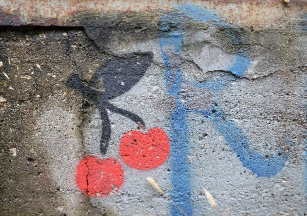 close up of a stencil graffiti of 2 cherries.  Beside the cherries is the letter R in blue.  Both are on concrete with a crack in it