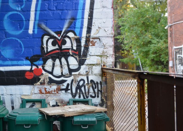 A silly face with big teeth with the word crush under it, graffiti on a wall in the alley. Green garbage bins in front of the wall.