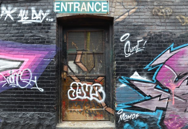 An old wood & glass door that has been painted over with graffiti.  A green and white sign saying Entrance is above the door.  Graffiti is on both sides of the door. 