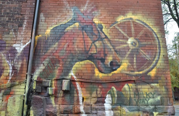 graffiti picture on a brick wall of a horse's head, a wagon wheel and spilt milk pouring from a large container. 
