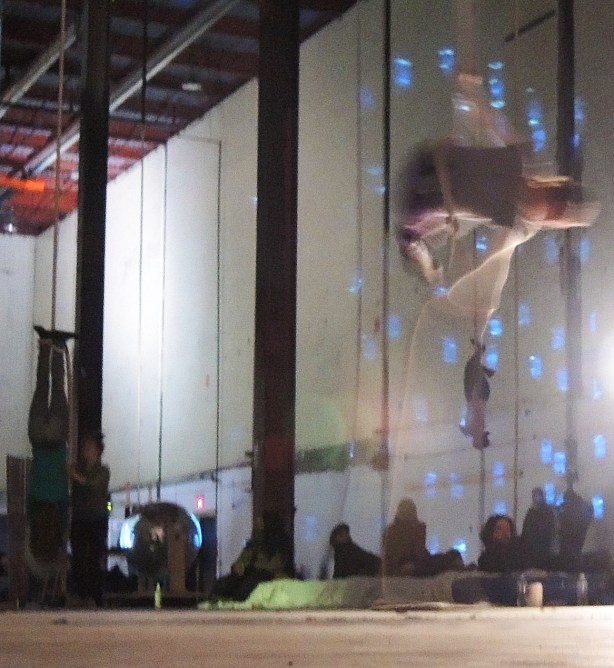 Three people are performing aerial movements on ropes hanging from the ceiling in a large dimly lit space 