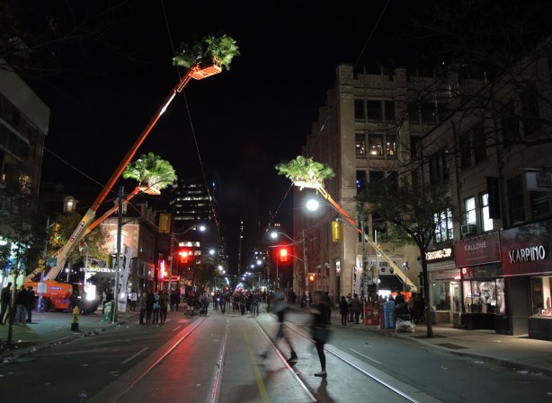 people on a street downtown at night.  3 large orange cranes with many small palm trees in their baskets overhand the street.  they are well lit from below. 