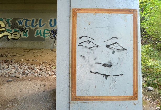graffiti under a bridge - yellowish gold frame with large line drawing of a smiling person. 