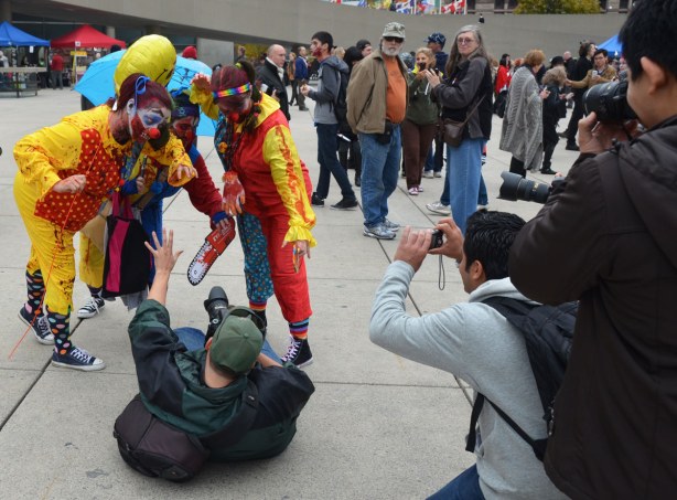 Three clown zombies are posed over a man who is lying on his back on the ground and holding a camera.  There is a small crowd around them, many with cameras.