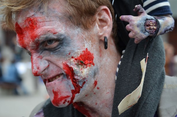 zombie man sneering.  there is a small hand behind his ear that is dangling from a fake zombie baby he is carrying on his shoulders (but that you can't see much of in this photo)