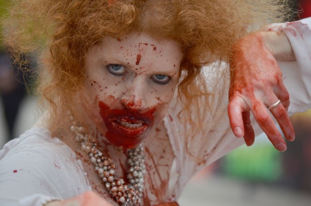 pale zombie with curly lught auburn hair, bloody chin and white dress.  She is glaring at the camera with her hand poised to reach for it. 