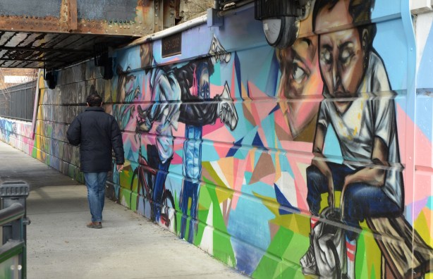 A man in a blue winter coat is walking under a bridge, beside a mural that is painted on the wall of the bridge. 