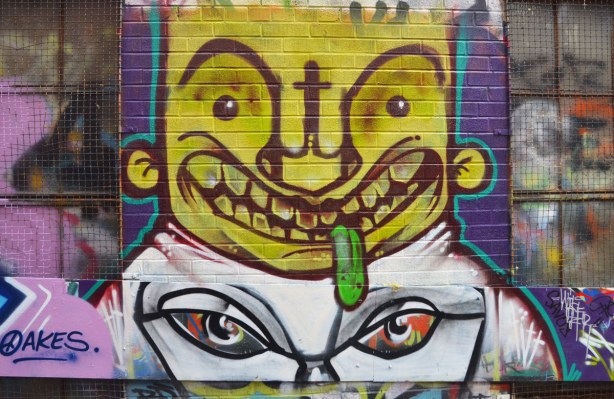 Street art in Graffiti Alley, picture of a man's face.  He seems to be leering with his tongue (which is green) hanging out of his mouth.  He is above another picture of a pair of anser eyes. 