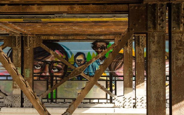 part of a mural beside the walkway under a railway bridge - taken from across the street,  The picture is of 4 people who seem to be looking at the camera. 