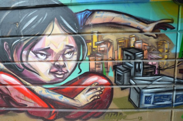part of a mural beside the walkway under a railway bridge - a woman sitting cross legged on the ground beside a miniature city