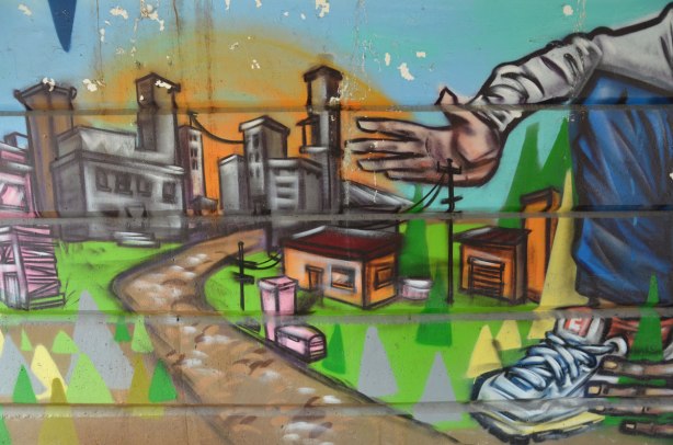part of a mural beside the walkway under a railway bridge - a man's hand reaching towards a miniature city.  roads, houses, tall buildings and hydro wires