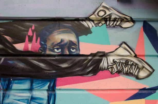 part of a mural beside the walkway under a railway bridge -  a pair of legs with sneakers on flying past a man's head. 