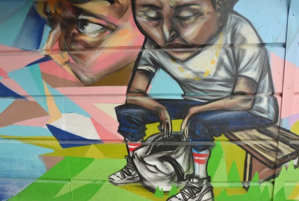 A man sitting on a bench who is looking down into a bag that is on the ground.  A face in profile is beside him