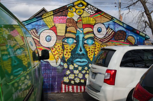 A van and car parked in front of a street art mural of a face in blue, yellow and red that is surrounded with stylized food, especially candy and popcorn. The neck is an iconic red and white striped popcorn container. The picture is reflected in the windows of the van.