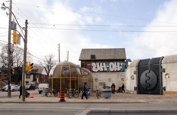At an intersection, looking across the street to a glass dome like structure that is a subway station entrance. Beside it is a building with a tag that says Uh Oh