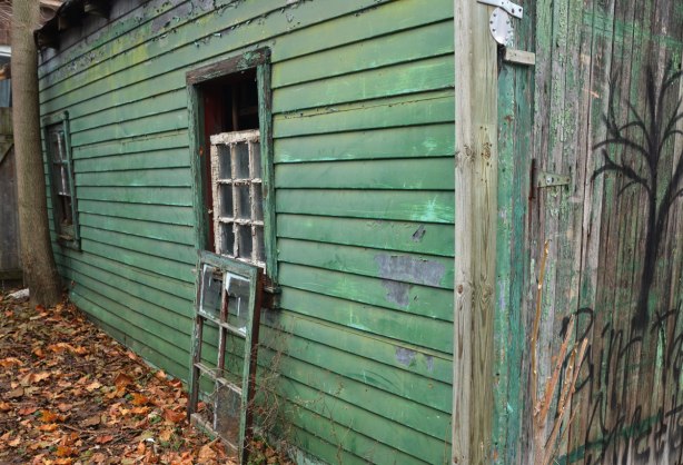 A green garage in need of paint.  A window frame is sitting on the ground beneath the window.  Dead leaves on the ground. 