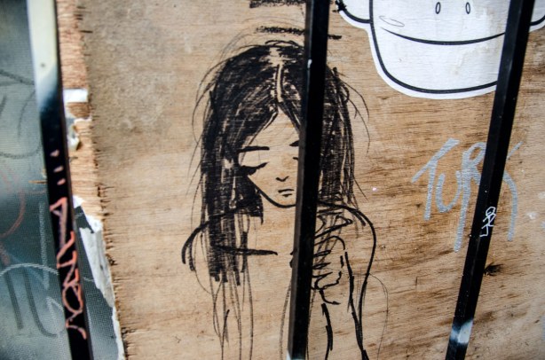A girl with long dark hair and with her eyes closed.  From the shoulders up with one arm and hand included in the picture. Drawn on plywood and behind bars. 