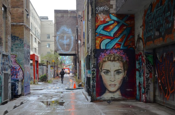looking down the alley, graffiti on the walls on both sides including a large woman's head by the artist Kairo.  She has curly hair and her face is about 7 feet tall. 