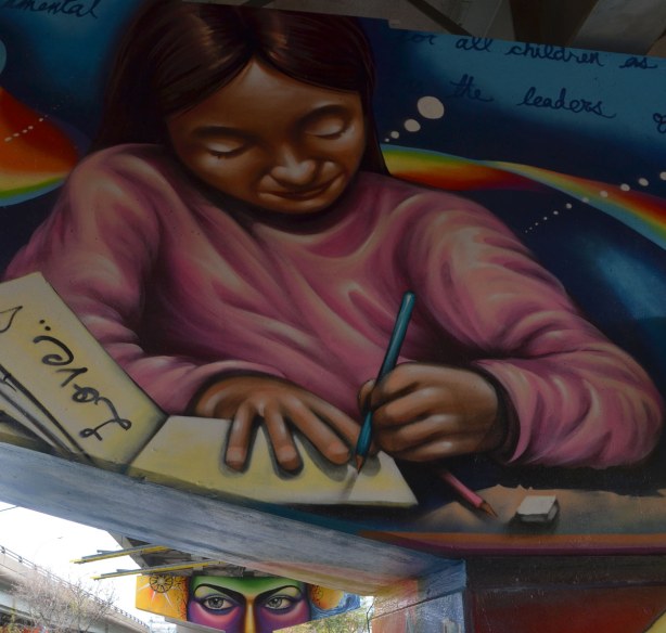 Paintings on the large T shaped concrete supports of the overpass, a young girl in a pink top sitting at a table and writing in a notebook.  The word love is written on one of the pages.  