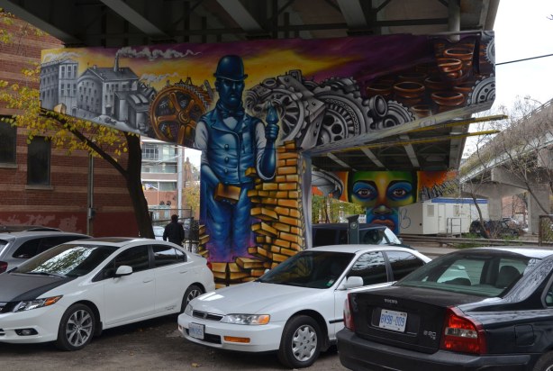 Mural on concrete pillar of a blue brick layer surrounded by symbols of industry such as factories, gears, beer bottles, bricks.  There is a parking lot surrounding the concrete support so there are cars parked in front of the pillar.  