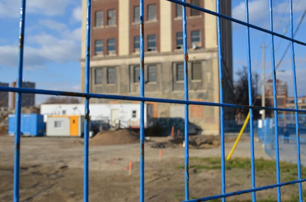 blue construction fence is in focus in the foreground with a construction site behind it.  A tall brick building stands in the construction site. 