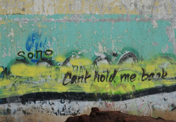 graffiti on the back of the Tower Automotive building, a tall brick building - the words cant hold me back are written in black on top of yellow and turquoise. 